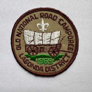 Może przedstawiać: Okrągła, haftowana naszywka z motywem wozu krytego. Naszywka zawiera napis "OLD NATIONAL ROAD CAMPOREE LAGONDA DISTRICT 1969". Obramowanie jest brązowe, a tło beżowe. Naszywka jest okrągła.