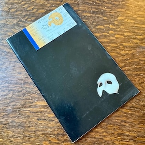 May include: A black theatre programme for "The Phantom of the Opera" with a white mask. A ticket is attached, displaying the date, time, and venue of the performance at the Pantages Theatre.