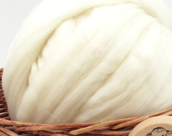 May include: A large, off-white bundle of wool roving, tightly packed and textured, fills the frame. The wool is nestled in a woven basket with a wooden accent. The natural fibers suggest a craft or textile product.