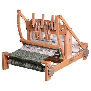 May include: A wooden table loom featuring a natural light wood frame with multiple vertical levers at the top. The loom is threaded with white warp yarns and has a partially woven fabric with a green and black herringbone pattern on the front beam.