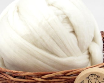 May include: A large, off-white bundle of wool roving, tightly coiled and filling a woven basket. The wool has a soft, fluffy texture, ideal for various fiber crafts. A wooden disc with a sheep illustration is visible.