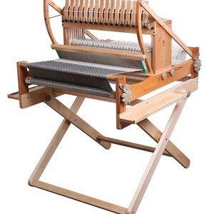 May include: A wooden weaving loom with a natural finish. The loom features a gray and white woven textile in progress. The frame has a folding stand and various wooden components for weaving. The loom is designed for textile creation.