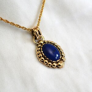 May include: A gold-toned necklace featuring an oval, deep blue pendant. The pendant is framed by a beaded border and hangs from a delicate chain. The necklace is displayed on a white surface.