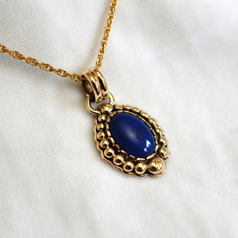 May include: A gold-toned necklace featuring an oval, deep blue pendant. The pendant is framed by a beaded border and hangs from a delicate chain. The necklace is displayed on a white surface.