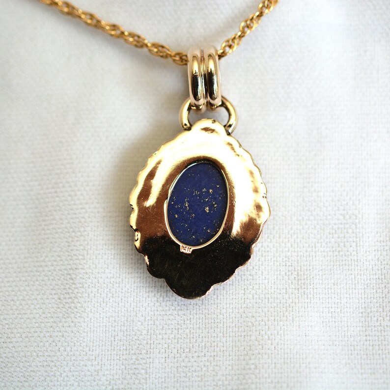 May include: A gold-colored pendant necklace with a dark blue oval stone. The pendant has a decorative, scalloped edge and is suspended from a delicate gold chain. The stone has flecks of gold within it.