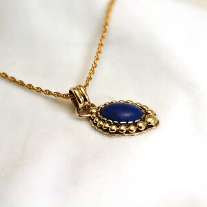 May include: A gold-colored necklace with a pendant featuring a deep blue oval stone. The pendant is surrounded by a beaded border. The chain is a delicate, twisted rope style.