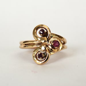 May include: A gold ring with three red gemstones set in a swirling, abstract design.