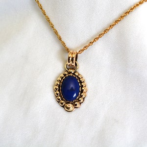 May include: Gold-toned necklace with an oval, deep blue pendant. The pendant is framed by a beaded border and hangs from a delicate chain. The necklace is displayed against a white background, highlighting the jewelry's design.