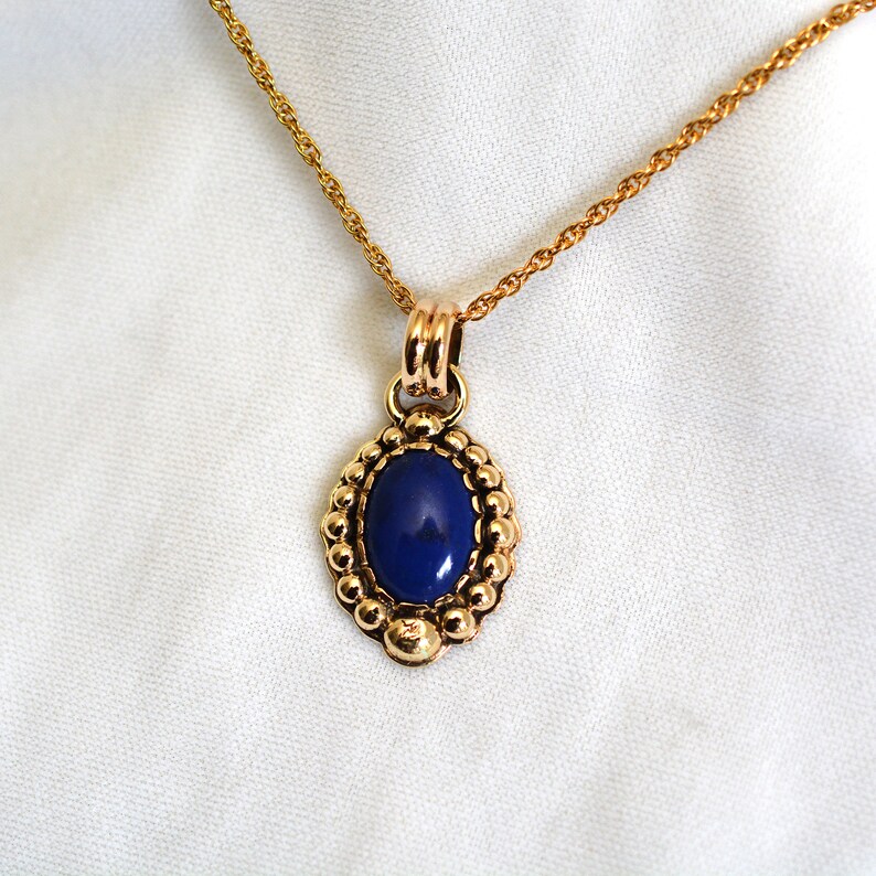 May include: Gold-toned necklace with an oval, deep blue pendant. The pendant is framed by a beaded border and hangs from a delicate chain. The necklace is displayed against a white background, highlighting the jewelry's design.