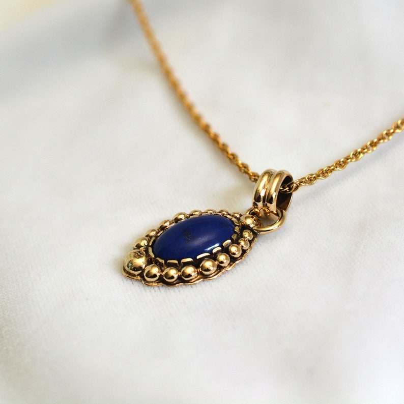 May include: A gold-colored necklace with a pendant featuring a deep blue oval stone. The pendant is surrounded by a beaded border and hangs from a delicate chain. The necklace is displayed on a white background.