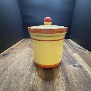 May include: A yellow ceramic jar with a lid. The jar has a cylindrical shape with a red and brown stripe around the top and bottom. The lid has a small red knob. The jar is sitting on a wooden surface.
