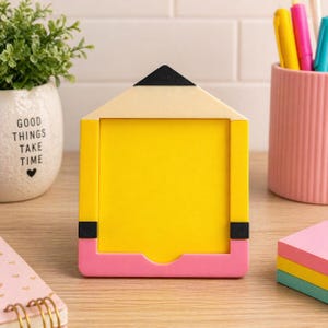 May include: A yellow, pink, and black pencil-shaped picture frame. The frame has a yellow center, a pink base, and a black tip. The frame is on a wooden surface, with a small plant and a pen holder in the background.