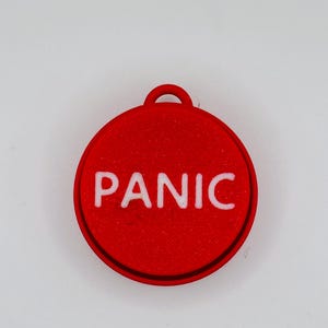 May include: A red, circular pendant with the word "PANIC" in white, bold letters. The pendant has a small loop at the top for attaching to a keychain or necklace. The design is simple and eye-catching.