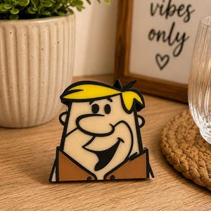 May include: A flat, rubberized character charm featuring a cartoon figure with bright yellow hair, a tan face, and a brown shirt. The design has bold black outlines, a wide smiling mouth, and a distinct nose, capturing the head and shoulders of the character.