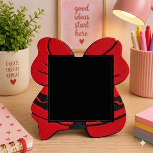 May include: A red and black bow-shaped picture frame with a black center. The frame is on a wooden surface, with a small potted plant and a pink desk lamp in the background. The frame has a unique design.