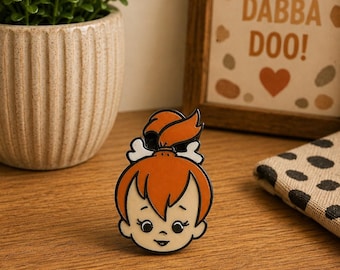Puede incluir: Un pin de esmalte metálico que muestra la cabeza de un personaje de dibujos animados con cabello naranja peinado en una coleta alta con un accesorio de hueso blanco. El personaje tiene tez color crema y una pequeña sonrisa.
