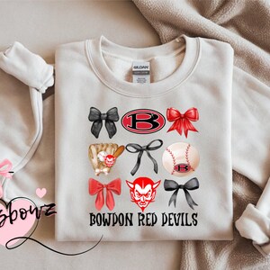 May include: Cream-colored sweatshirt featuring a design with bows, a baseball, and a devil mascot. The text "BOWDON RED DEVILS" is printed below the graphics. The sweatshirt has a crew neck and long sleeves.