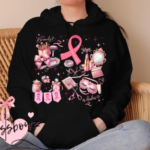 May include: Black hoodie with a pink ribbon and beauty-themed graphics. The design includes makeup, scissors, and the words "Beauty", "Hope", and "lashes". The hoodie features a drawstring hood and a kangaroo pocket.