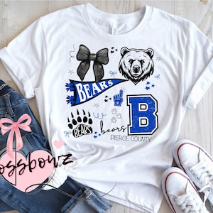 Op de afbeelding: Wit T-shirt met een blauw en zwart grafisch ontwerp met een beer, strik, pootafdrukken en het woord "Bears". De letter "B" staat in een blauwe lijst met de woorden "Pierce County".