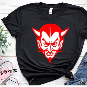 May include: A black short-sleeve t-shirt featuring a bold, centered graphic of a red and white devil face with pointed horns, a sharp goatee, and an intense expression. The shirt has a crew neckline and rolled sleeves, presenting a clean, graphic design.