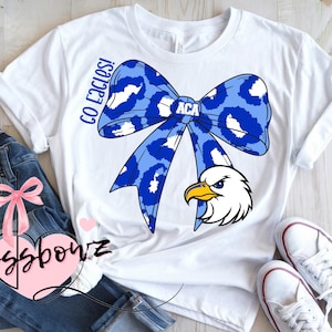 May include: White t-shirt featuring a large blue bow with a leopard print design and the text "ACA". An eagle head graphic is below the bow. The words "Go Eagles!" are printed vertically on the left side of the bow.