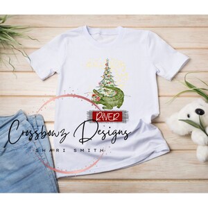K&ouml;nnte beinhalten: Wei&szlig;es T-Shirt mit einem gr&uuml;nen Alligator, der einen Weihnachtsbaum mit roten Kugeln h&auml;lt. Der Alligator sitzt auf einem roten Schild mit der Aufschrift "River". Das Shirt tr&auml;gt den Text "Crossbowz Designs Shari Smith" unten.