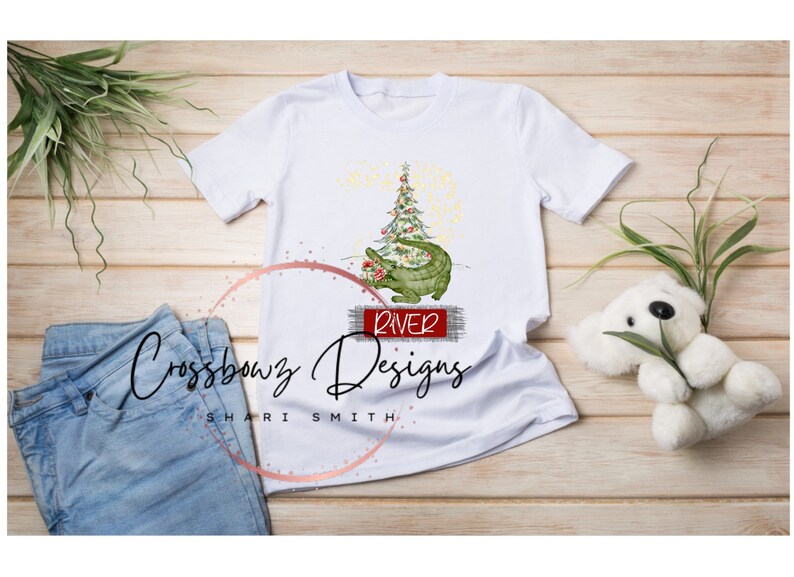 K&ouml;nnte beinhalten: Wei&szlig;es T-Shirt mit einem gr&uuml;nen Alligator, der einen Weihnachtsbaum mit roten Kugeln h&auml;lt. Der Alligator sitzt auf einem roten Schild mit der Aufschrift "River". Das Shirt tr&auml;gt den Text "Crossbowz Designs Shari Smith" unten.