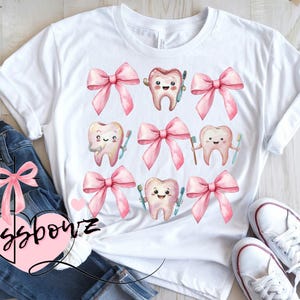 May include: White t-shirt featuring a repeating pattern of smiling cartoon teeth with toothbrushes and pink bows. The text "Crossbowz" is visible on the bottom left of the image. The shirt is paired with blue jeans and white sneakers.