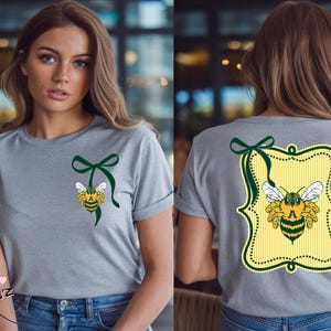 May include: A heather gray t-shirt featuring a bee design with a green bow. The bee is yellow, black, and white. The back of the shirt has a similar bee design inside a yellow and green frame.