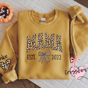May include: Mustard yellow sweatshirt with the word "MAMA" in leopard print lettering. Below the word "MAMA" is the word "EST." and the number "2022". The left sleeve has the name "Chloe" and a leopard print heart. A pumpkin-shaped bowl is in the upper left corner.