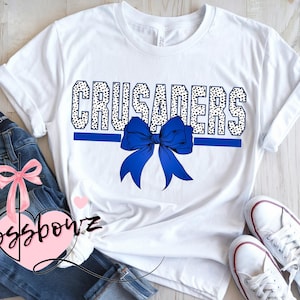 May include: White t-shirt with the word "CRUSADERS" in a black and white polka dot design, a blue bow, and a blue horizontal stripe. The shirt is paired with blue jeans and white sneakers.