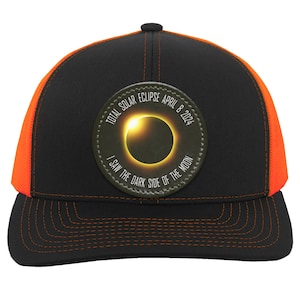 May include: Black and orange trucker hat with a circular patch that reads "Total Solar Eclipse April 8, 2024 I Saw The Dark Side Of The Moon" and a graphic of a solar eclipse.