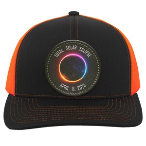 May include: Black and orange trucker hat with a circular patch that reads "Total Solar Eclipse April 8, 2024".