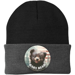 May include: A black and grey beanie hat with a circular badge. The badge shows a baby bear illustration set against an American flag and forest scene. The words "BABY BEAR" are in white.