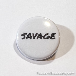 May include: A white button with the word "SAVAGE" in bold, black letters. The button is round and has a glossy finish. The button is a pinback button.