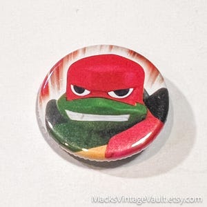 May include: A round button featuring a cartoon image of Raphael from Teenage Mutant Ninja Turtles. The button has a red mask, green face, and white teeth. The background has red and white stripes. The text "MacksVintageVault.etsy.com" is at the bottom.