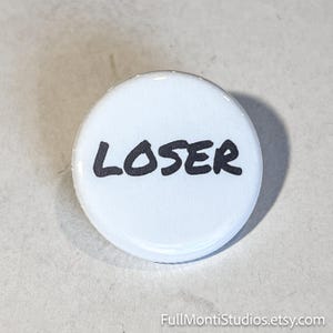 May include: A white button with the word "LOSER" in bold, black letters. The button is round and has a glossy finish. The button is a novelty item.