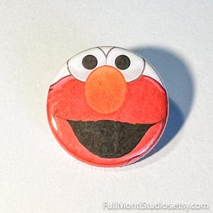 May include: A round button featuring a cartoon character's face. The character has a red face, a large orange nose, black eyes, and a wide black mouth. The button has a glossy finish and is set against a white background.