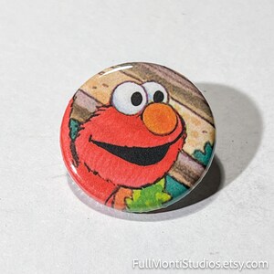 May include: A round button featuring the Sesame Street character Elmo. The button has a glossy finish and a colorful design, with Elmo's red face, orange nose, and black smile. The background has a wooden plank design. The text "FullMontiStudios.etsy.com" is at the bottom.