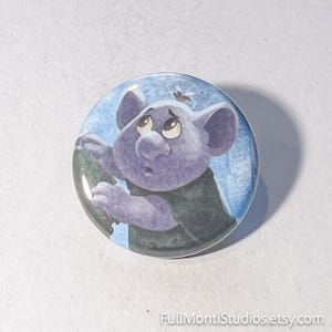 May include: A round button featuring a cartoon illustration of a purple troll-like creature with large ears and a sad expression. The background is blue with white accents. The button has a glossy finish.