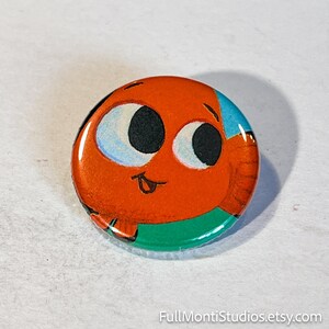 May include: A round button with a cartoon fish design. The fish is orange with large black eyes, blue accents, and a green bottom. The button has a glossy finish and the text "FullMontiStudios.etsy.com" at the bottom.
