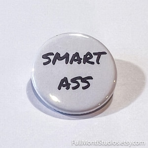 May include: A round, white button with the words "SMART ASS" in black, handwritten-style lettering. The button has a glossy finish and a simple, bold design. The button is a novelty item.