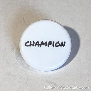 May include: A white button with the word "CHAMPION" in black, bold, handwritten-style font. The button is round and has a glossy finish. The button is a motivational item.