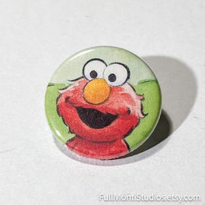 May include: A round button featuring a smiling cartoon character with red fur, a large orange nose, and big white eyes. The background is light green with darker green accents. The button is from FullMontiStudios.etsy.com.