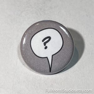 May include: A round, gray button with a white speech bubble containing a black question mark. The button has a glossy finish. The text "FullMontiStudios.etsy.com" is at the bottom. This button is a fun accessory.