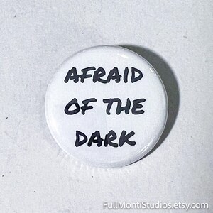 May include: A white button with the phrase "AFRAID OF THE DARK" in black, bold letters. The button is round and has a glossy finish. The Etsy shop name "FullMontiStudios.etsy.com" is at the bottom.
