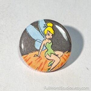 May include: A round button featuring a cartoon illustration of Tinkerbell. The fairy is depicted with blonde hair, a green dress, and blue wings, set against a dark background. The button has a glossy finish and is likely a collectible item.