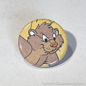 May include: Round button with a cartoon bear illustration. The bear is brown with a light brown belly and a red nose, set against a yellow background. The button has a glossy finish and is likely a pin or badge.