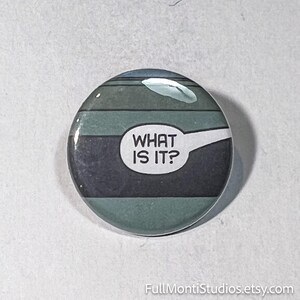 May include: Round button with a comic book style design. The button features a speech bubble with the text "WHAT IS IT?" in white. The background has horizontal stripes in shades of blue and green.