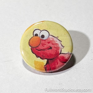 May include: A round button featuring a watercolor illustration of Elmo from Sesame Street. The button has a yellow background and a glossy finish. The text "FullMontiStudios.etsy.com" is at the bottom.